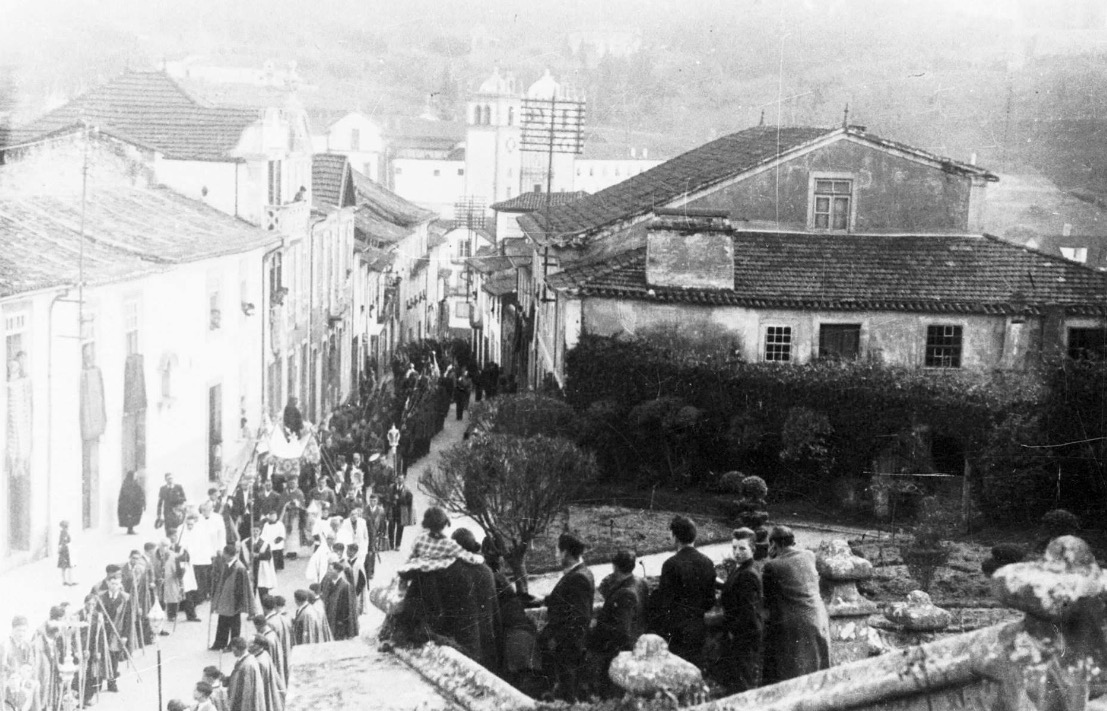Fotografia antiga a preto e branco da rua e da casa, com uma procissão a passar
